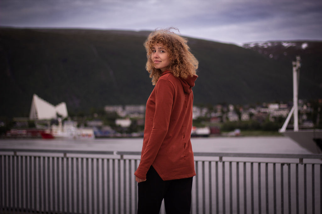A woman with curly light brown hair stands on a bridge or walkway by the water, wearing a rust-orange hooded sweatshirt and black pants, with a cityscape and green hills in the background.