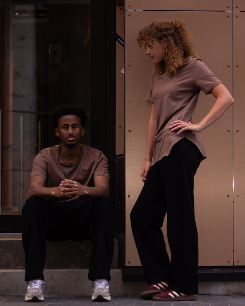 Two models posing in front of a modern building facade with warm-toned metal panels. One model is seated on the steps wearing a brown t-shirt and black pants, while the other stands wearing a taupe t-shirt and black pants.