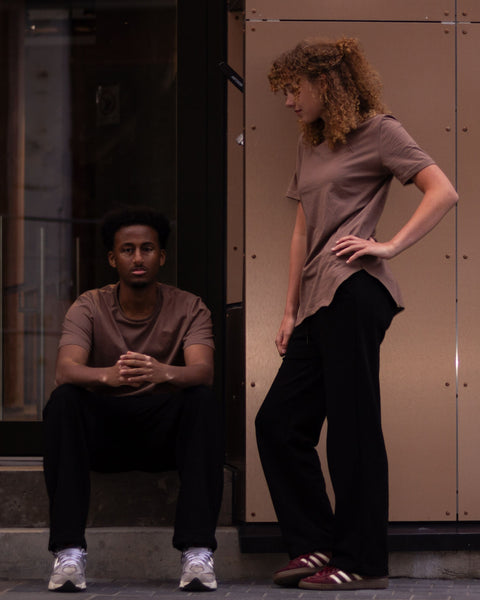 Two models posing in front of a modern building facade with warm-toned metal panels. One model is seated on the steps wearing a brown t-shirt and black pants, while the other stands wearing a taupe t-shirt and black pants.