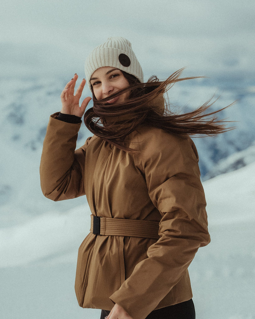 Woman in a brown coat and Icon soft sand beanie standing in a snowy landscape