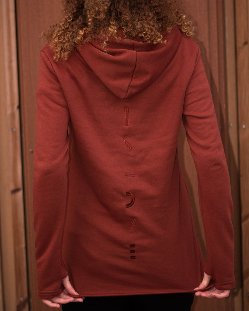 A person viewed from behind wearing a rust-colored hooded long-sleeve top with decorative dark embroidery down the back, standing against a wooden door.