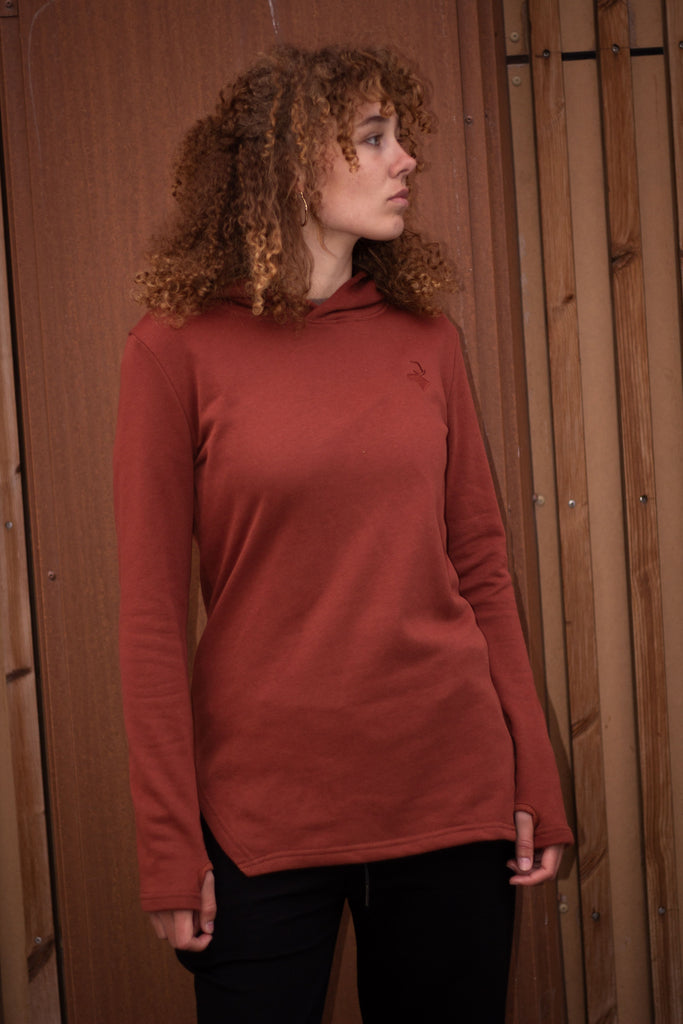 A woman with curly brown hair wearing a rust-colored long-sleeve mock neck top and black pants, standing in front of a wooden slatted wall.