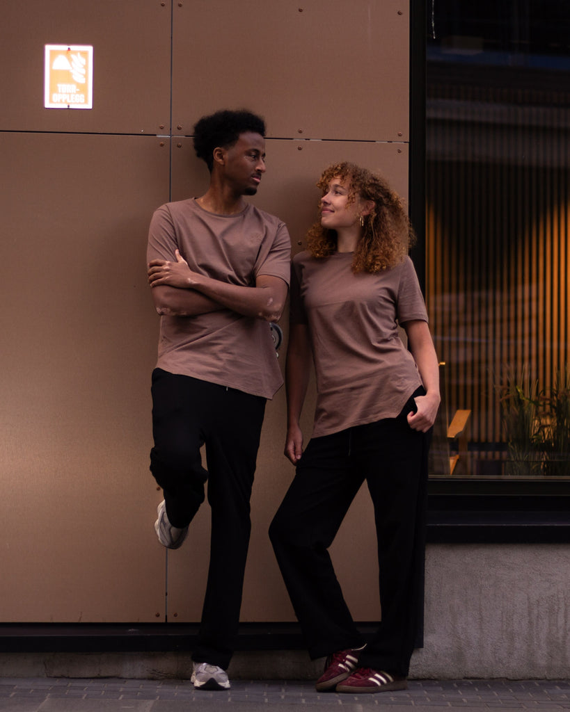 Two people standing outdoors against a tan building wall, wearing matching muted mauve-brown short-sleeve T-shirts and dark pants.