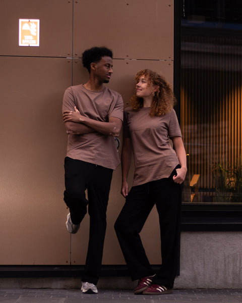 Two models posing in front of a modern building facade with warm-toned metal panels. One model is seated on the steps wearing a brown t-shirt and black pants, while the other stands wearing a taupe t-shirt and black pants.