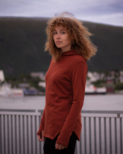 A woman with curly light brown hair stands outdoors by a railing, wearing a rust-orange long-sleeve mock-neck top and dark pants, with water, boats, and a mountain in the background.