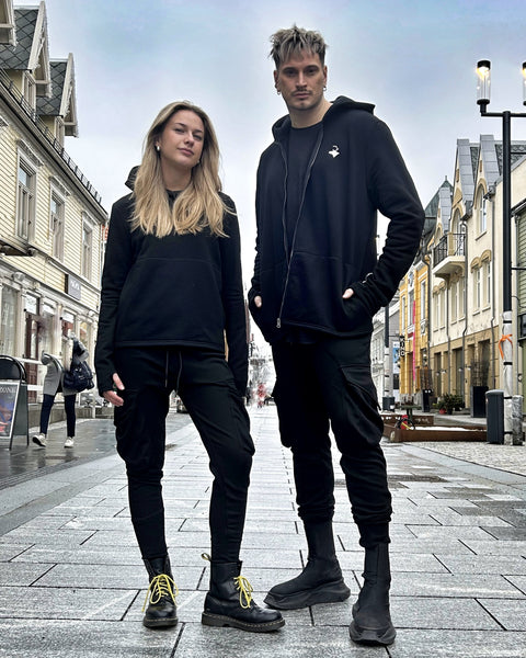 Benjamin and Signe standing side by side on the Tromsø high-street next to Rein Love Brand Store wearing black cargo pants and black sweat shirts. The male model is on the right and the female model is on the left.