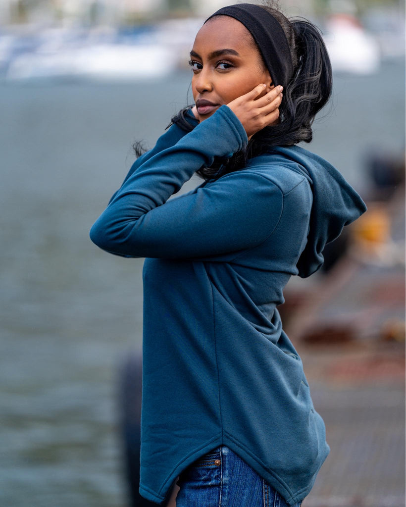 She is wearing the petrol blue hoodie Identity runner with the slogan 'Don’t follow the herd, Stay Wild' on the sleeves, paired with jeans, standing near a body of water.