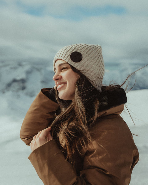 Woman in a brown coat and Icon soft sand beanie standing in a snowy landscape